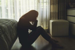 Eine Frau sitzt auf dem Boden vor ihrem Bett, in einem dunklen Schlafzimmer und stützt ihren Kopf mit ihren Händen