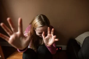 eine Frau sitzt mit dem Rücken zur Wand auf dem Boden und hält ihre Hände abwehrend in die Luft
