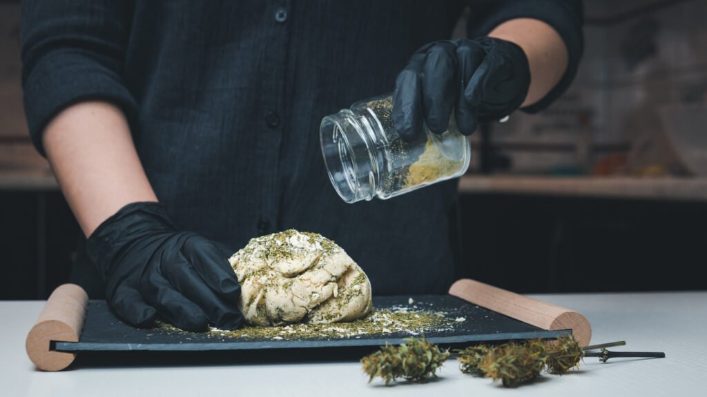 Hier sieht man eine Köchin mit schwarzen Handschuhen, die in einen Brotteig Cannabis gibt. Sie betreibt also infused Cooking.