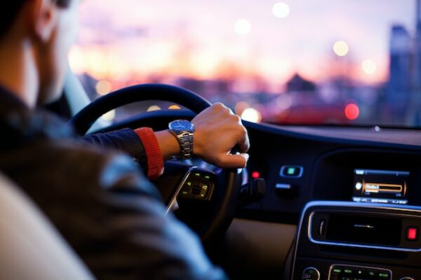 Hier ist ein junger Mann zu sehen, wie er mit einer Hand das Steuer eines Autos hält. Eine allgemeine Verkehrskontrolle kann auf der Straße jederzeit stattfinden.