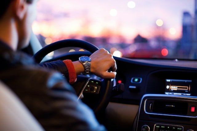 Hier ist ein junger Mann zu sehen, wie er mit einer Hand das Steuer eines Autos hält. Eine allgemeine Verkehrskontrolle kann auf der Straße jederzeit stattfinden.