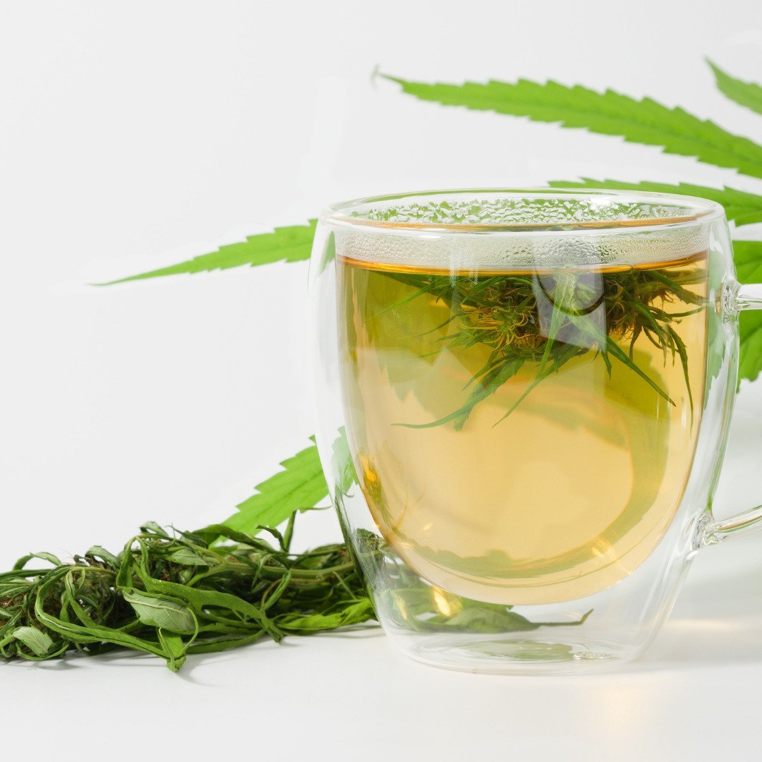Ein Teeglas mit Wasser ist zu sehen, in dem eine Cannabisblüte schwimmt. Also ein Cannabis-Tee. Daneben und dahinter sind Cannabisblätter zu erkennen.