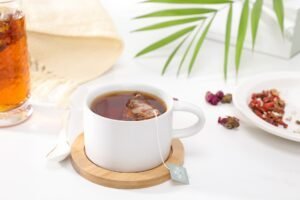 Eine weiße Tasse mit rotem Tee ist zu sehen. Aus der Tasse hängt die Schnur des Teebeutels. Schräg dahinter liegen rote Kräuter.