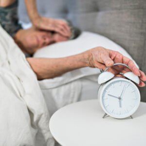 eine Frau liegt im Bett, mit der Hand am Kopf und schaut auf den Wecker, der 5 Uhr morgens zeigt.