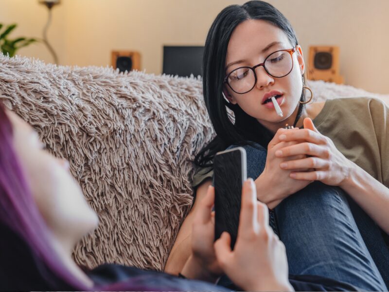 Cannabiskonsum bei Jugendlichen ist rückläufig. Hier sieht man zwei junge Frauen, eine mit einem Handy, eine mit einem Joint im Mund.