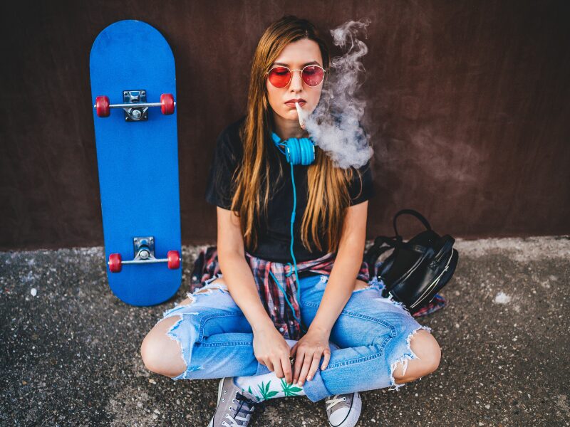 Prävention bei Jugendlichen! Ist diese zeitgemäßg und ausreichend? Ein junges Mädchen zieht an einem Joint, mit blauen Kopfhörern, einem blauen Skateboard und einer orangenen Sonnenbrille.